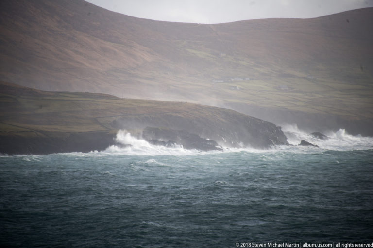 Large white waves crash against a dark rocky headland beneath rain-softened hills.