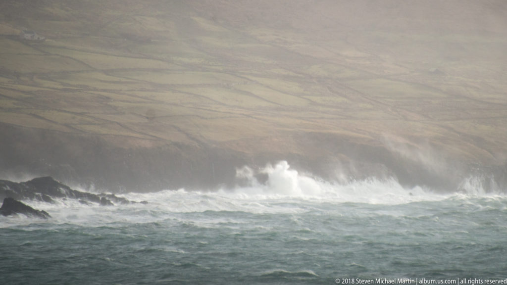 White surf bursts over a dark rock shelf in rough gray-green water, with misty hills behind.