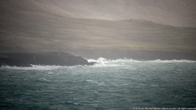 White waves break along a low dark cliff beneath pale green fields and misty hills.
