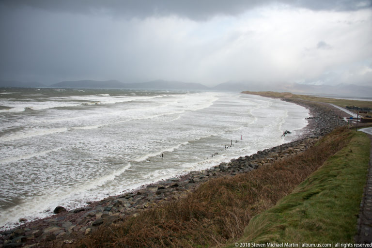 Multiple lines of white surf break along a broad gray bay beside a narrow coastal road and rocky embankment.