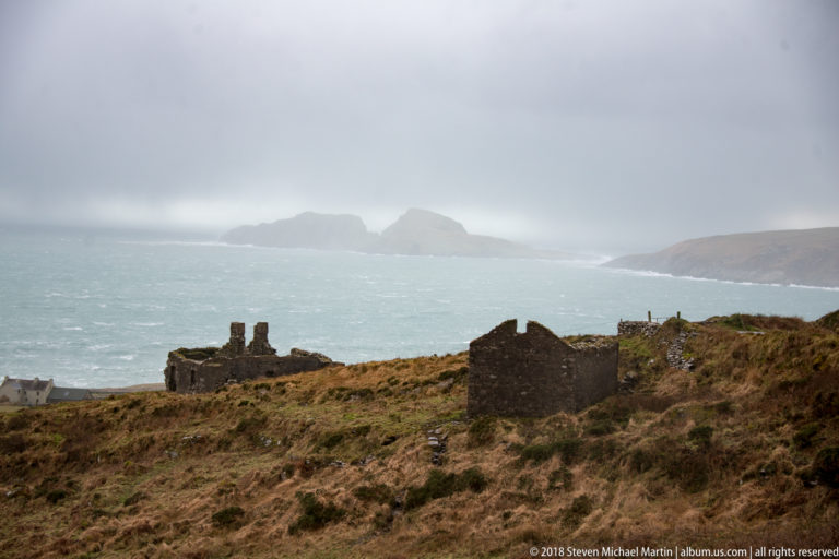 Roofless stone ruins on brown coastal grassland above a rough sea with islands in the distance.
