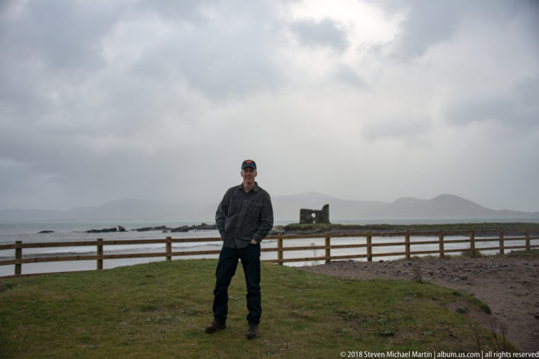 Man standing on grass by a wooden fence, with a small offshore stone ruin and misty hills behind.
