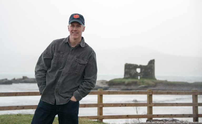 Waist-up portrait of a man in a dark cap, with a small offshore ruin and pale mountains behind.