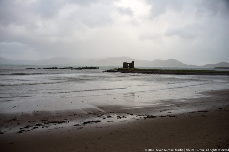 Wide wet beach facing a small offshore islet with a roofless stone ruin beneath gray clouds.