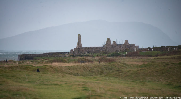 Large roofless stone complex with tall pointed gables on grassland below a hazy mountain.