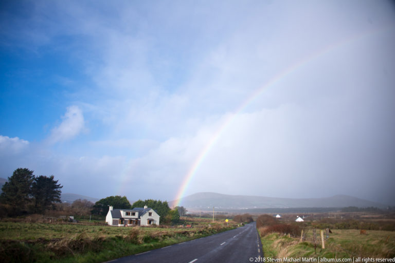 Rainbow arches over a straight rural road and white house, with blue sky to one side and rain cloud to the other.
