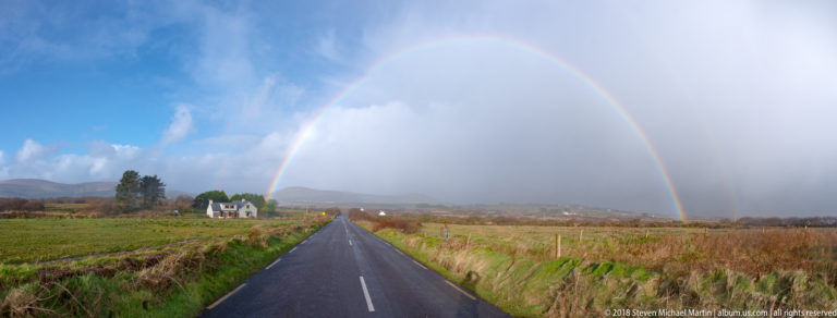 Full rainbow arches over a straight two-lane road through fields, with rain and blue sky divided across the horizon.