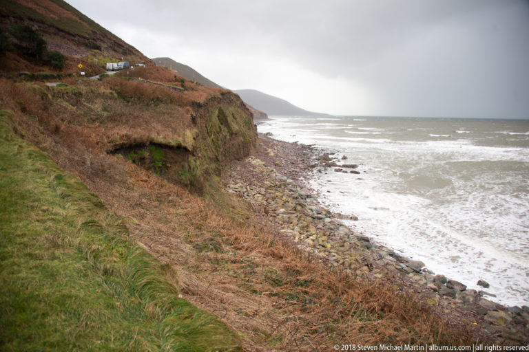 Grass-topped eroding cliff above a boulder beach and rough gray sea, with misty headlands beyond.
