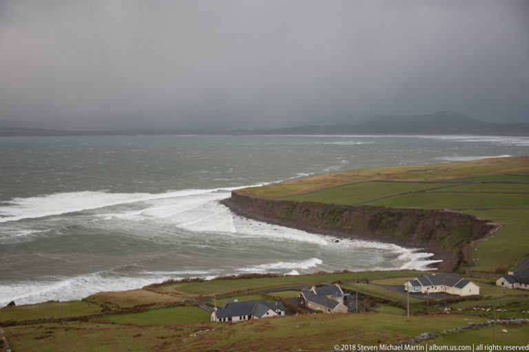 Small houses and green fields above a cliff-edged coast with white surf and dark rain beyond.