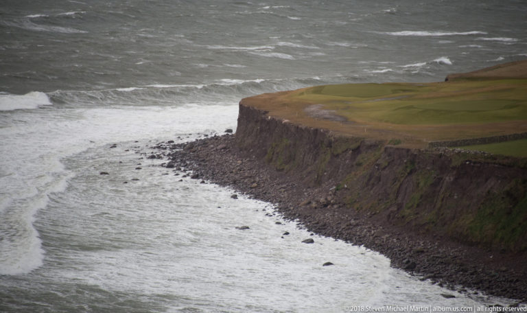 Green pasture ends at steep dark cliffs above a boulder shore and bands of white surf.