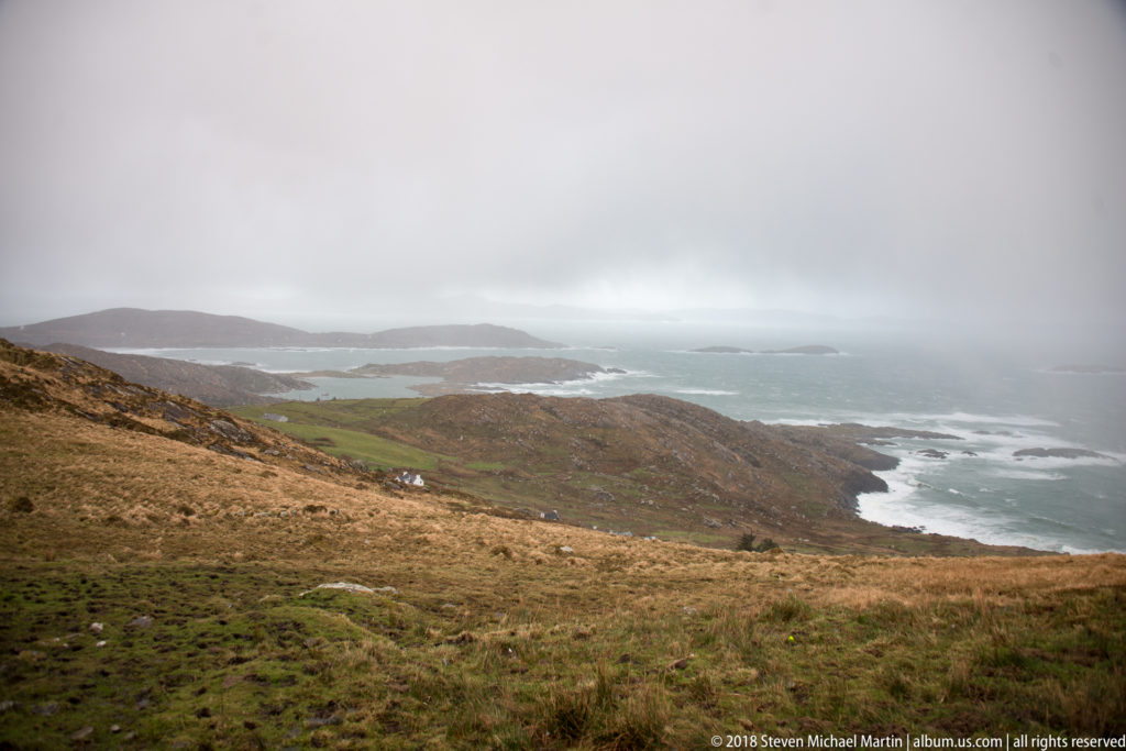 High view over tawny moorland toward rocky headlands, small islands, and gray Atlantic water in rain.
