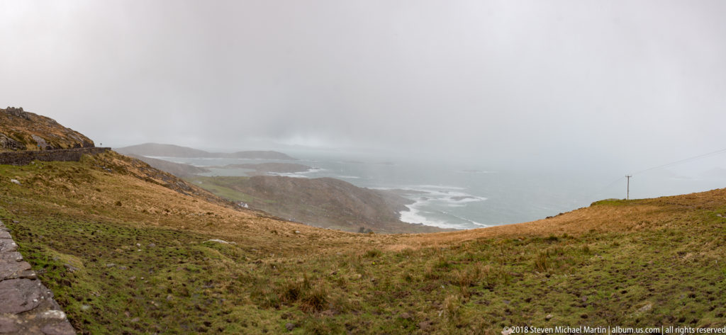 Narrow road on a steep grassy hillside above misty cliffs and rough Atlantic water.