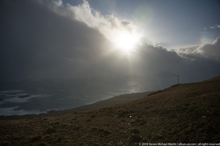 Sunlight bursts through dark storm clouds onto the Atlantic above a shadowed grassy slope.