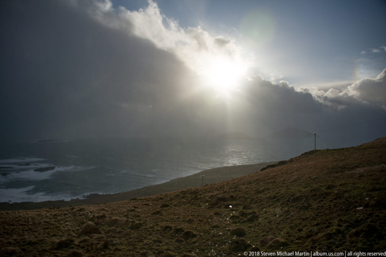Bright sun opening in dark clouds sends rays over gray ocean beyond a low grassy slope.