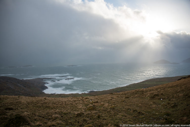 Sunbeams illuminate gray-blue Atlantic water beyond tawny grass and white surf along a rocky coast.
