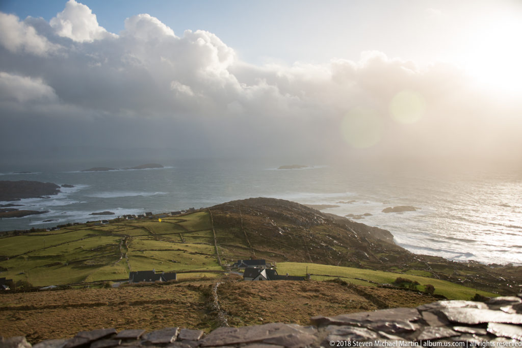 Sunlit Atlantic water beyond green patchwork fields, a rocky headland, and small offshore islets beneath heavy clouds.