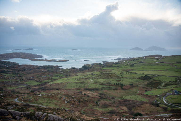 Patchwork green coastal fields descending toward rocky islands and blue Atlantic channels under towering rain clouds.