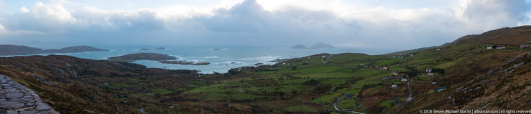 Steep green coastal valley descending to rocky Atlantic islets beneath a visible rain shower and dark mountain slopes.