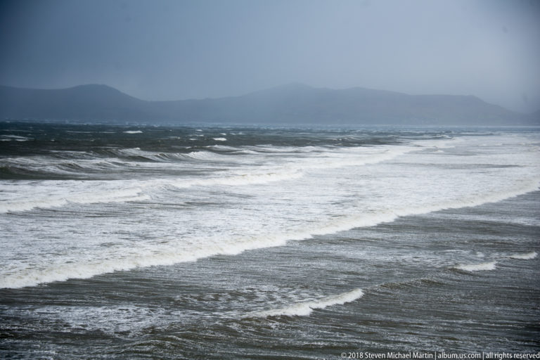 Rows of white-capped Atlantic waves beneath dark misty mountains and a heavy gray sky.
