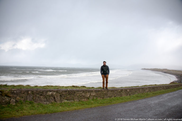 Person standing on a low roadside wall above a stormy bay with breaking waves and distant mist.
