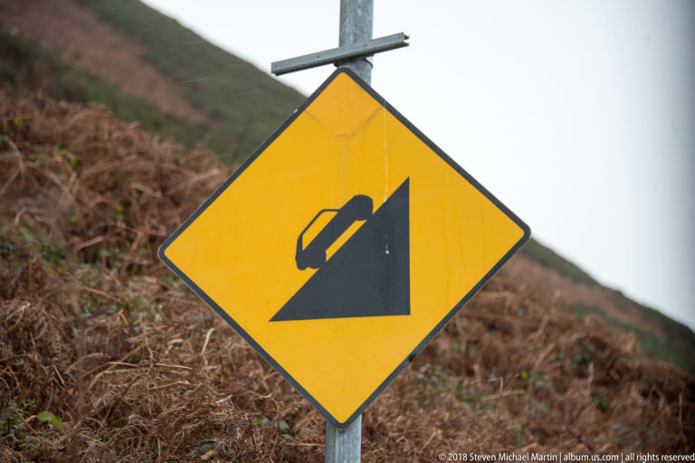 Yellow diamond road sign with a black car descending a steep slope, set against brown bracken.