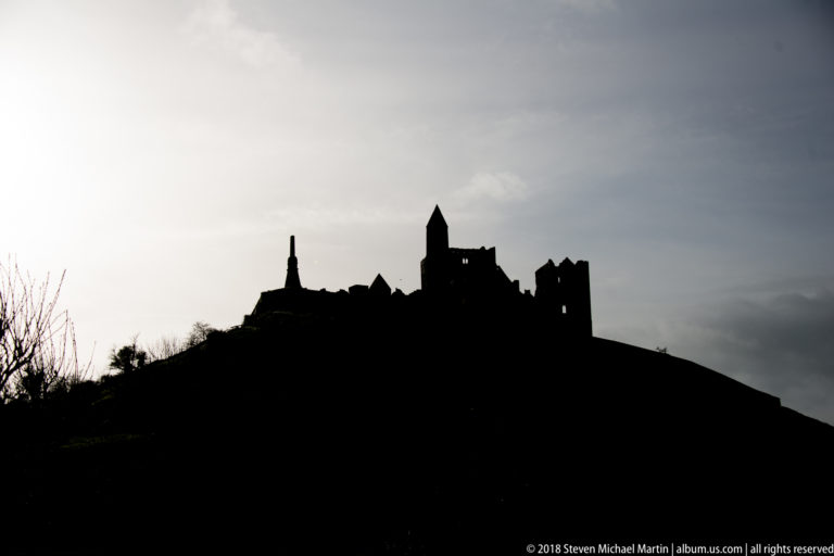 Dark silhouette of the Rock of Cashel complex crowning a hill beneath a pale overcast sky.