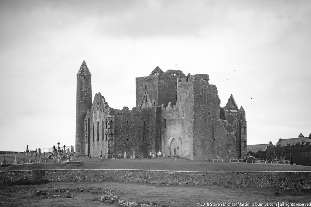 Monochrome view of Rock of Cashel ruins with round tower, large stone walls, grave markers, and small figures.
