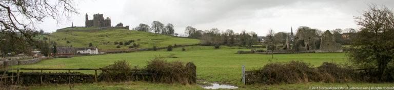 Ruined abbey walls in green fields with trees and the Rock of Cashel visible on the distant hill.