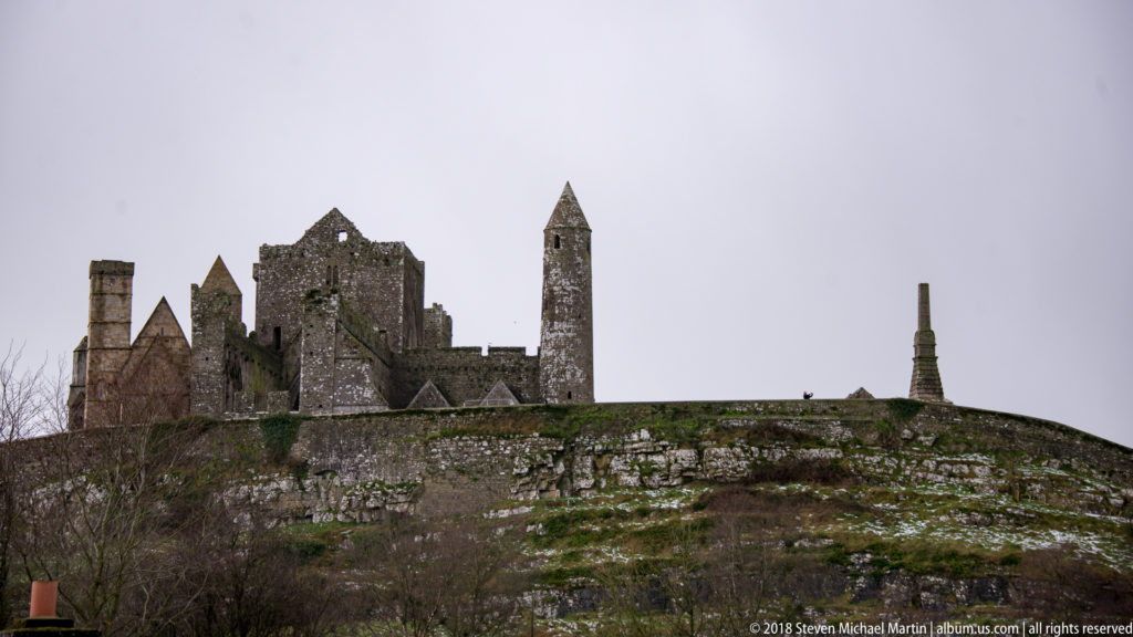Rock of Cashel towers and gables above a pale limestone escarpment, with a slender monument at right.