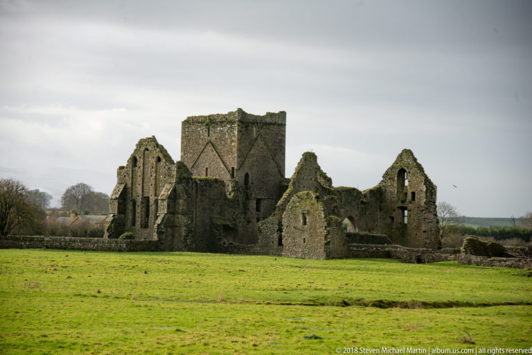 Roofless stone abbey ruins with a tall square central tower standing in a green pasture under gray skies.