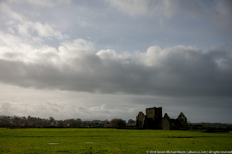 Distant medieval abbey ruins across green fields beneath a large bank of dark gray clouds.