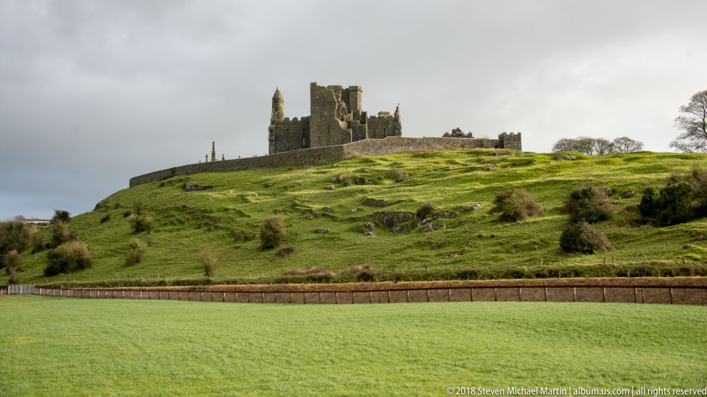Wide view of the Rock of Cashel on a green limestone ridge beyond a cultivated field.