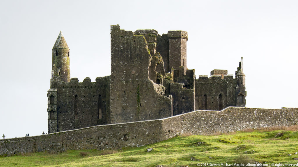 Close view of the Rock of Cashel with round tower, ruined stone walls, and perimeter wall above a grassy slope.