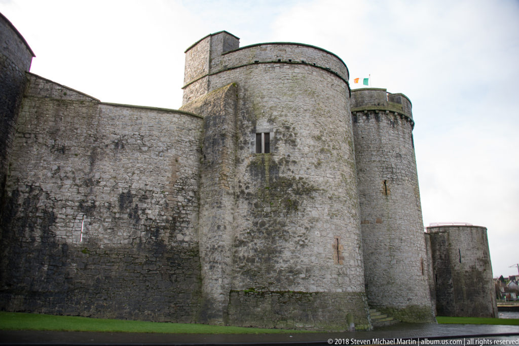 Round limestone towers and high curtain wall of King John’s Castle viewed from grass at close range.