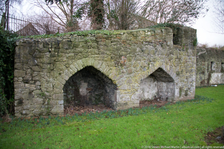 Low weathered stone wall with two adjoining arches standing beside grass and leafless trees.
