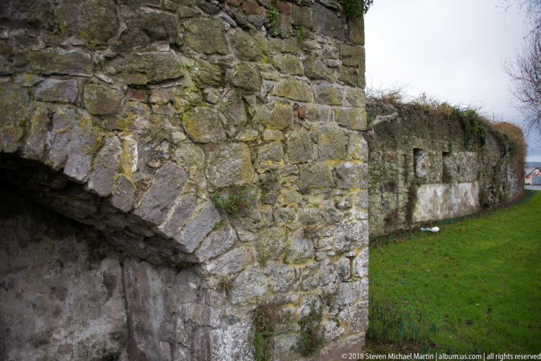 Rough stone wall with a broad masonry arch, patched surfaces, grass, and vegetation growing between the stones.