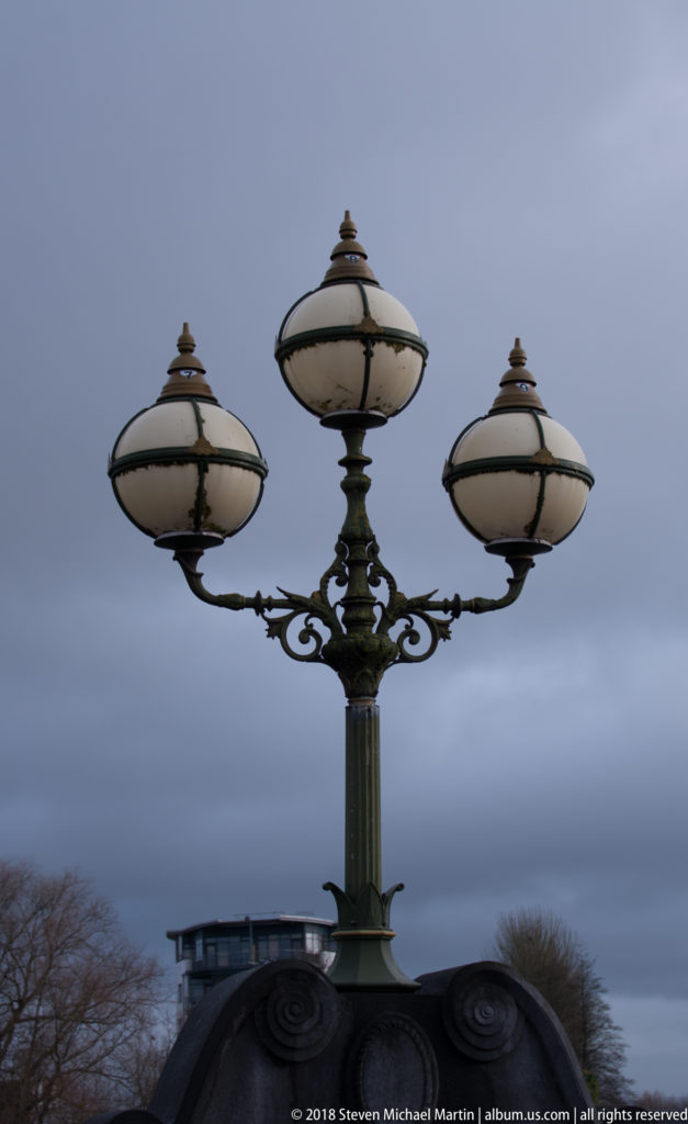 Ornate three-globe street lamp with scrolling metalwork silhouetted against an overcast sky near King John’s Castle.