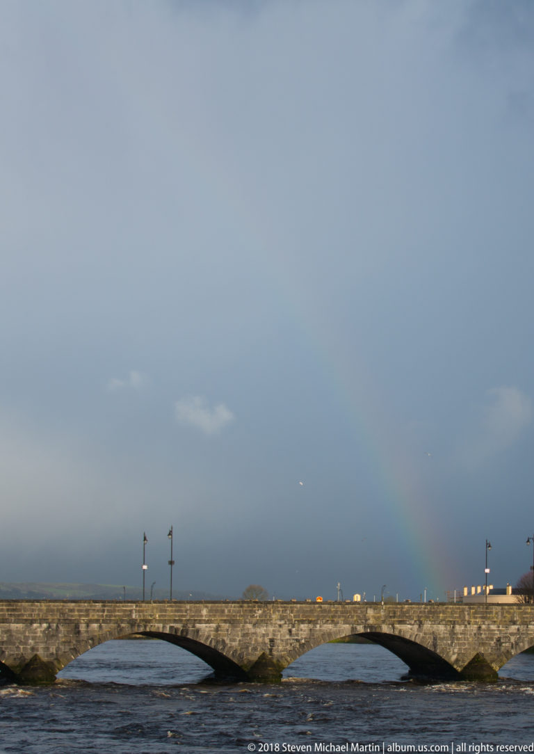 Faint rainbow over the low stone arches of Thomond Bridge and the River Shannon beneath a dark gray sky.
