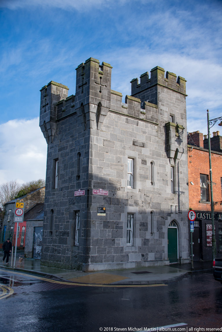Small gray stone building with crenellated parapets, corner projections, narrow windows, and a green door on a city street.