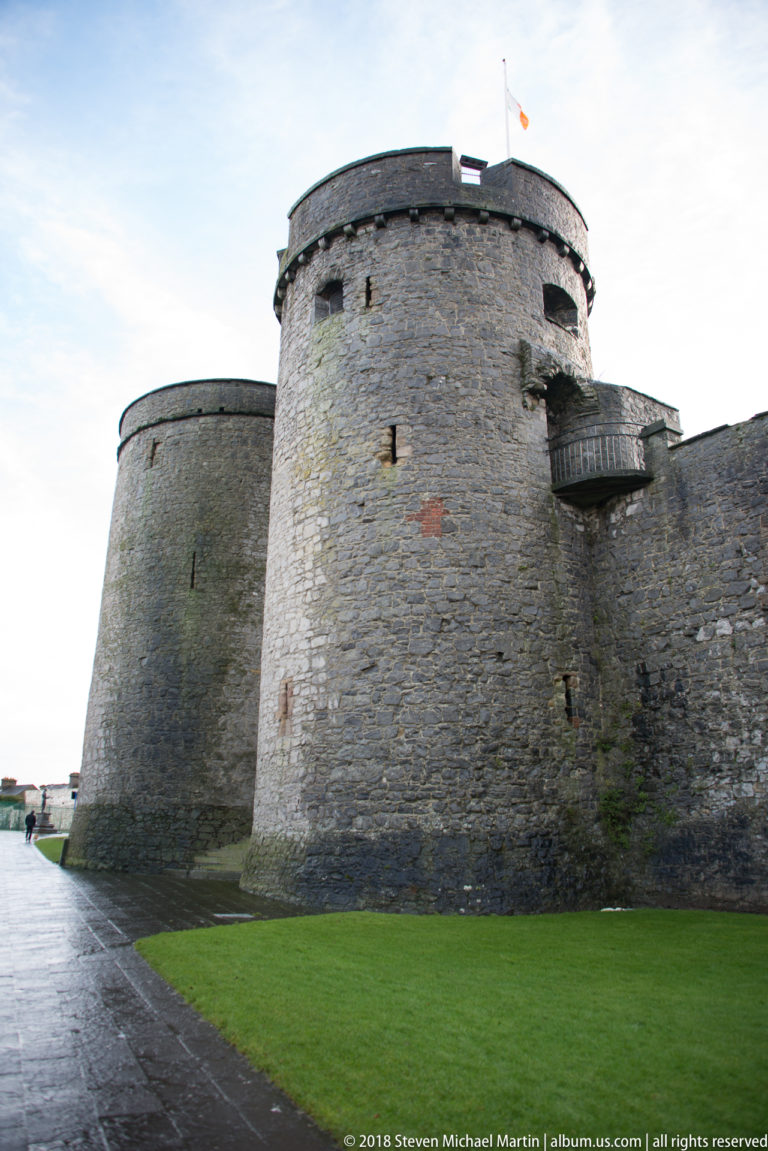 Curving stone towers and connecting defensive wall of King John’s Castle beside a wet path and green lawn.
