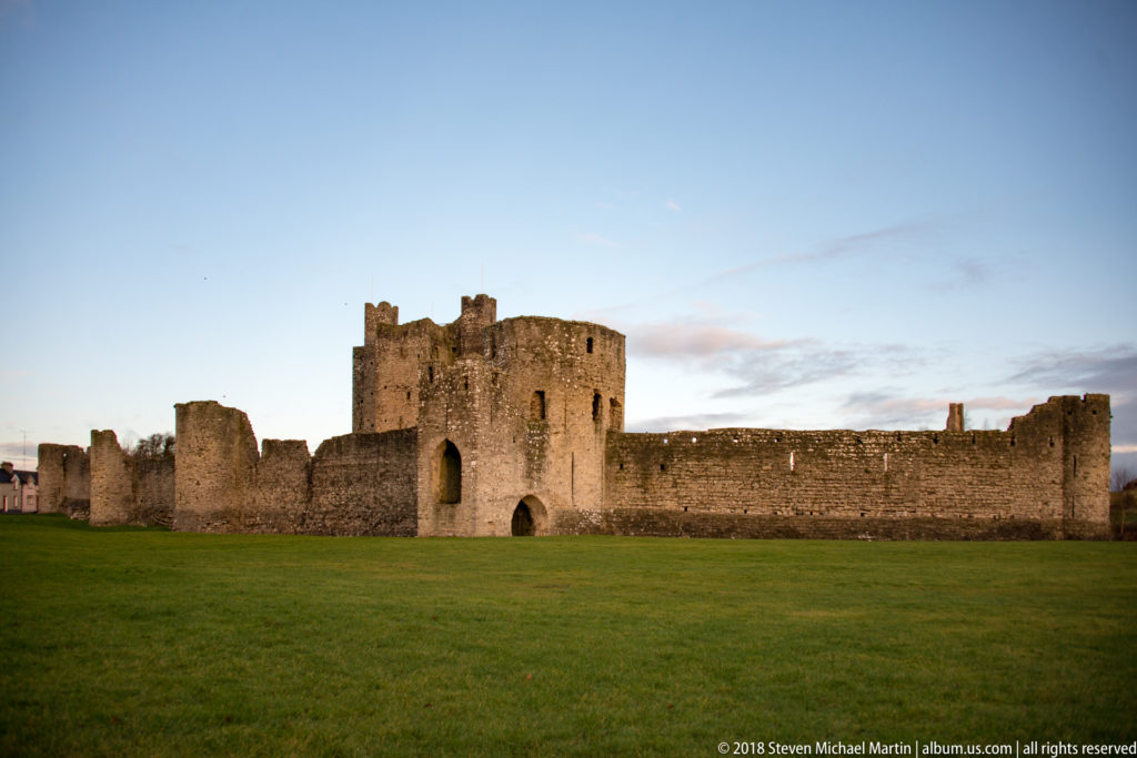 Long stone curtain wall and arched gate tower of Trim Castle, with the central keep rising behind across a broad green lawn.