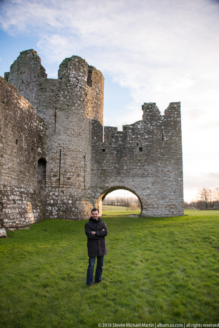 Visitor standing on green grass inside Trim Castle, with a rounded tower and broad arched wall opening behind.