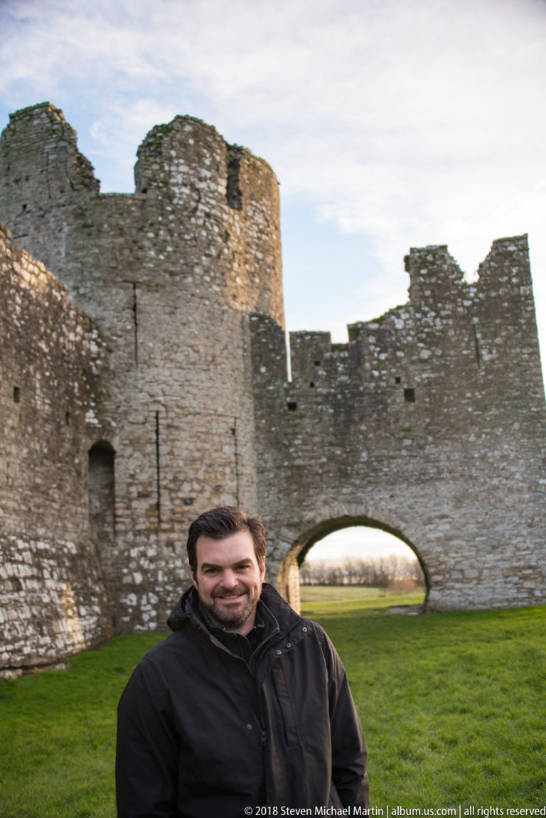 Smiling visitor in a dark jacket standing close to the camera, with Trim Castle’s stone tower, wall, and arch behind.