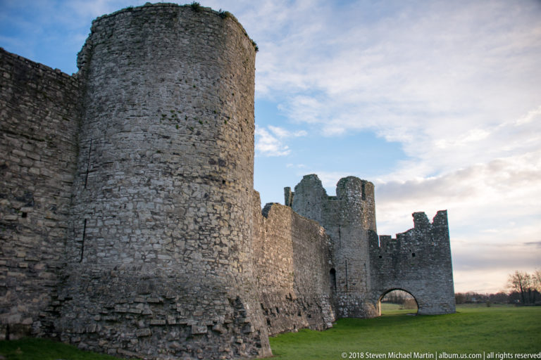 Curved stone tower at left leading along a long curtain wall toward additional towers and an arched opening in the distance.