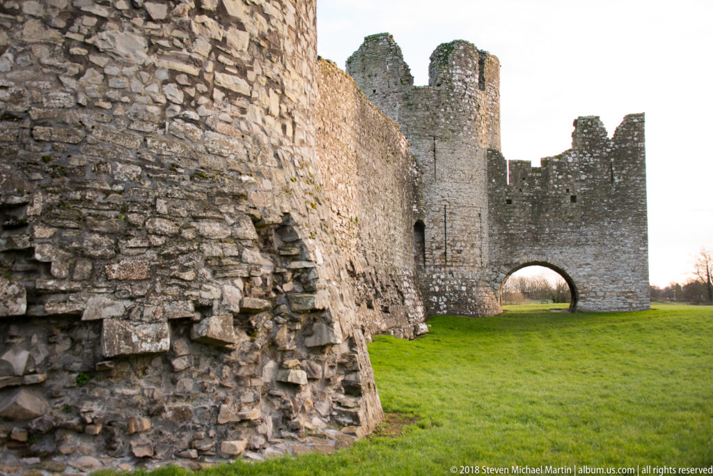 Close oblique view along rough Trim Castle masonry toward a square tower and broad arch, with bright grass beside the wall.