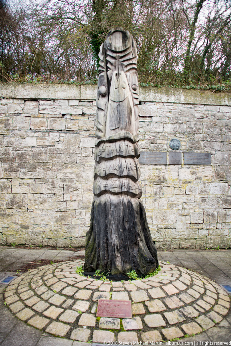 Large carved wooden figure in a robe standing on circular paving before a stone wall, with a small plaque at the base.