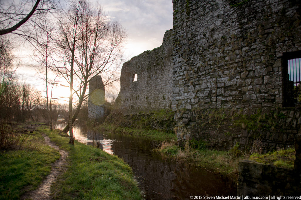 Long broken stone wall beside a narrow river channel and footpath, backlit by low sun through leafless trees.