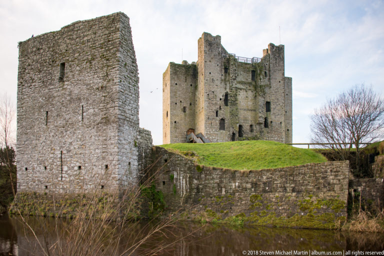 Trim Castle keep on a grassy rise behind a nearer stone tower and low wall beside a narrow watercourse.