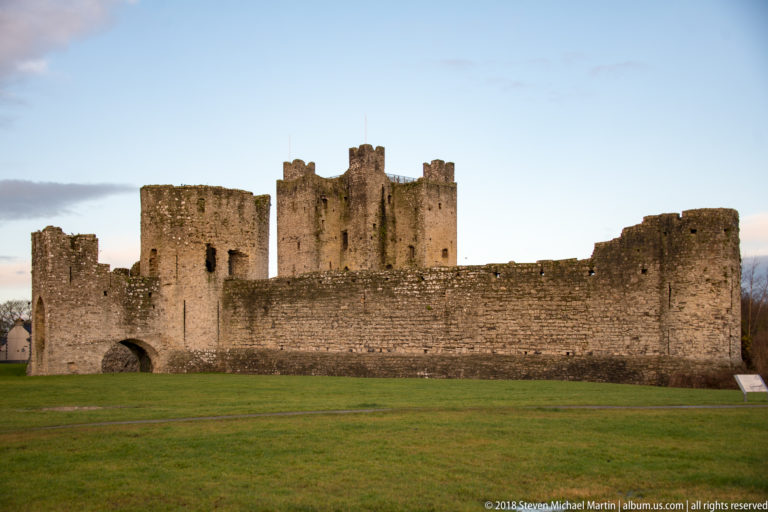 Central keep of Trim Castle rising behind a lower stone curtain wall with an arched opening and flanking defensive towers.