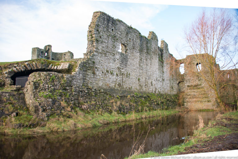 Roofless stone walls and broken gables beside calm river water, with collapsed masonry and leafless trees around the ruins.